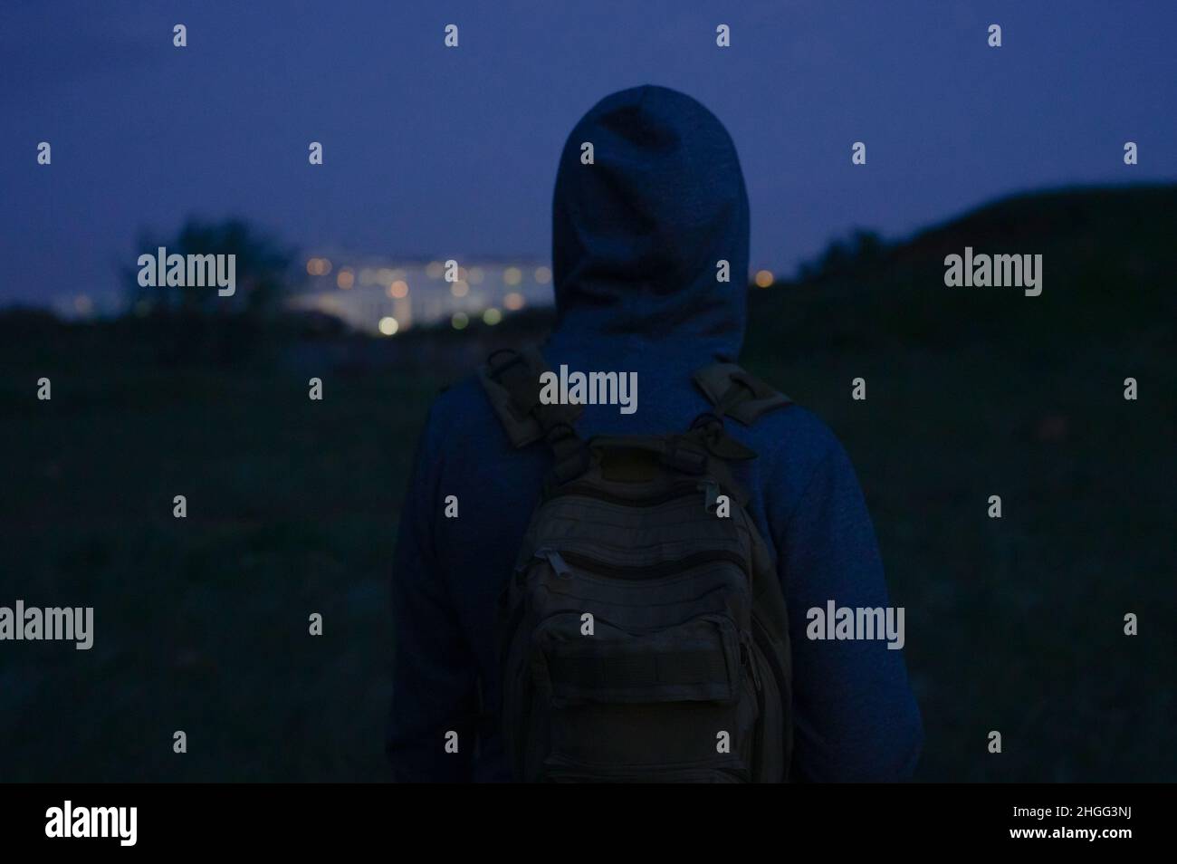 hombre sin hogar en hoodie con mochila mirando en los edificios de la ciudad de noche durante el senderismo al atardecer Foto de stock
