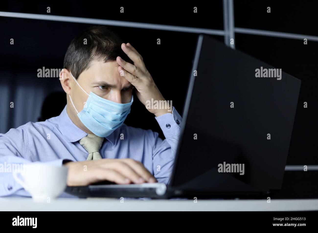 Hombre con máscara facial y ropa de oficina que mira enfocada a la pantalla del portátil. Concepto de resolución de una tarea difícil Foto de stock
