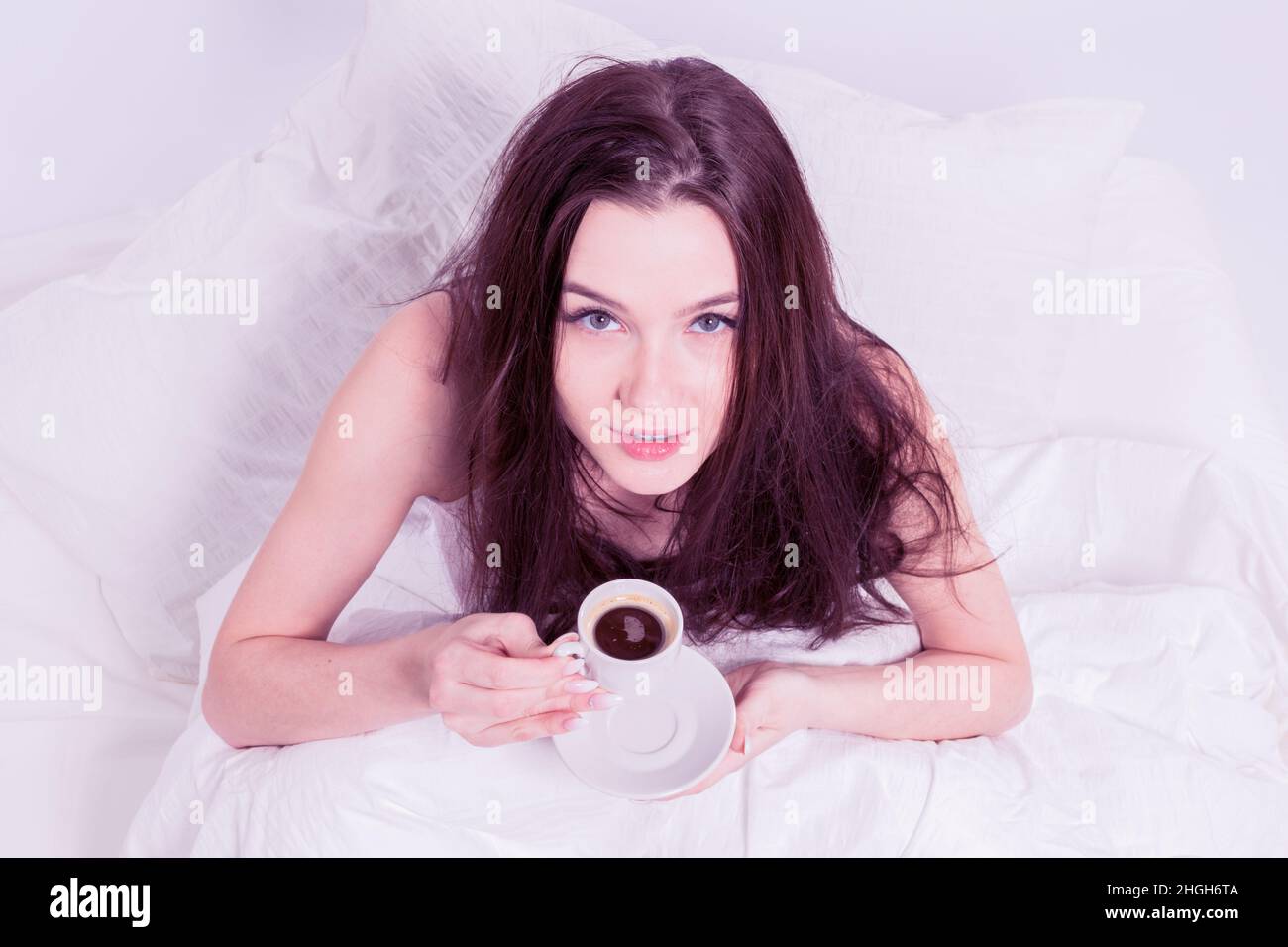 La mujer está sentada en la cama con café. Desayuno sabroso. La morena tiene una taza con una bebida caliente. Dormitorio blanco. Buenos días. Foto de stock