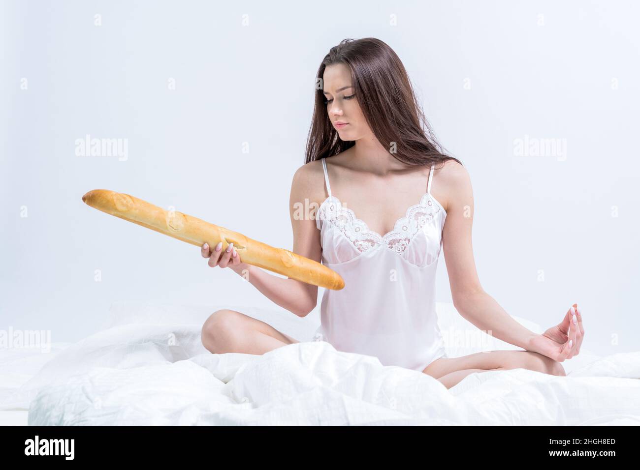 Una mujer está sentada en una cama blanca sosteniendo una baguette. La morena está tomando el desayuno con pan en la cama. Buenos días. Desayuno en el hotel. Foto de stock