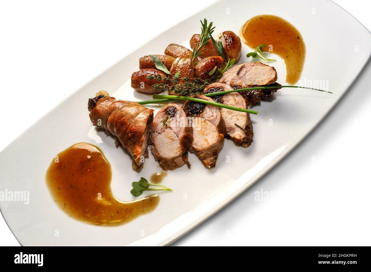 Rollo de conejo horneado relleno de ciruelas pasas, rodajas con patatas y hierbas aromáticas en plato blanco aislado sobre blanco Foto de stock