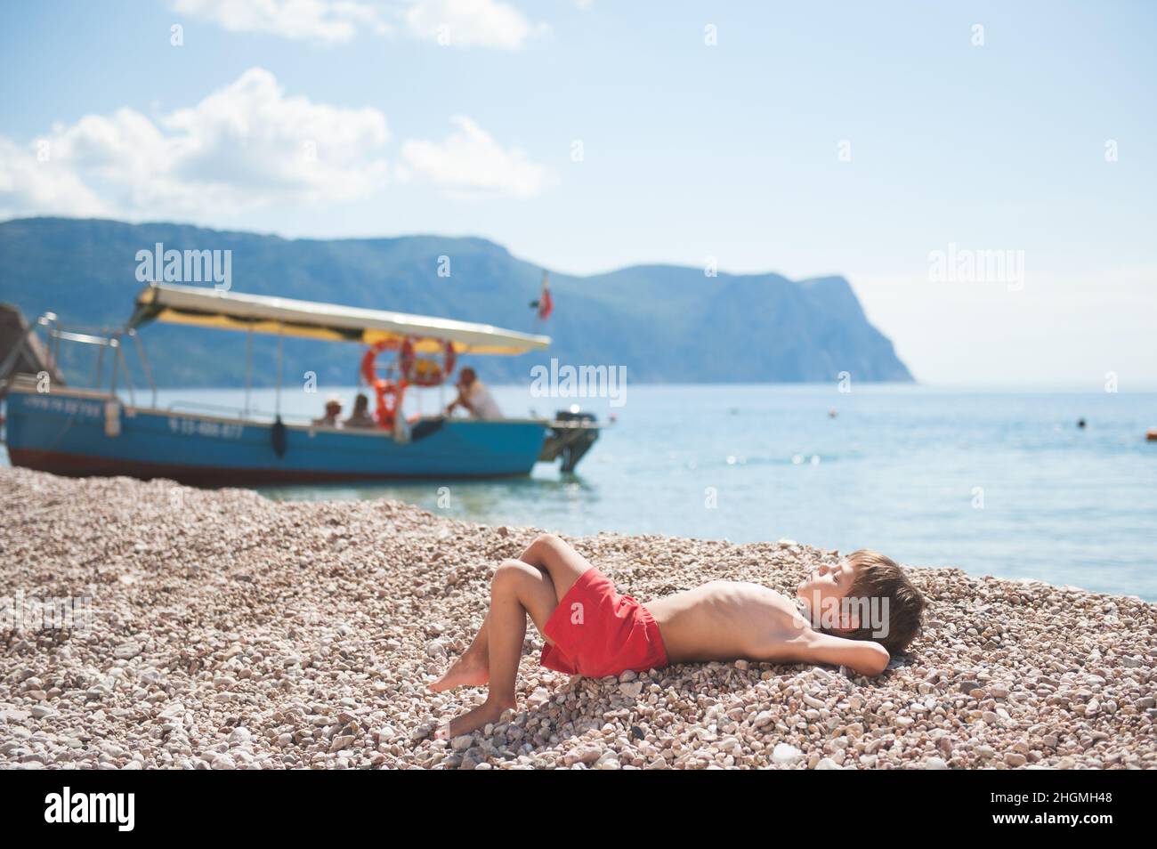 feliz sano soñando niño pequeño tumbado en la playa de verano con barco y montaña rocosa en el fondo Foto de stock