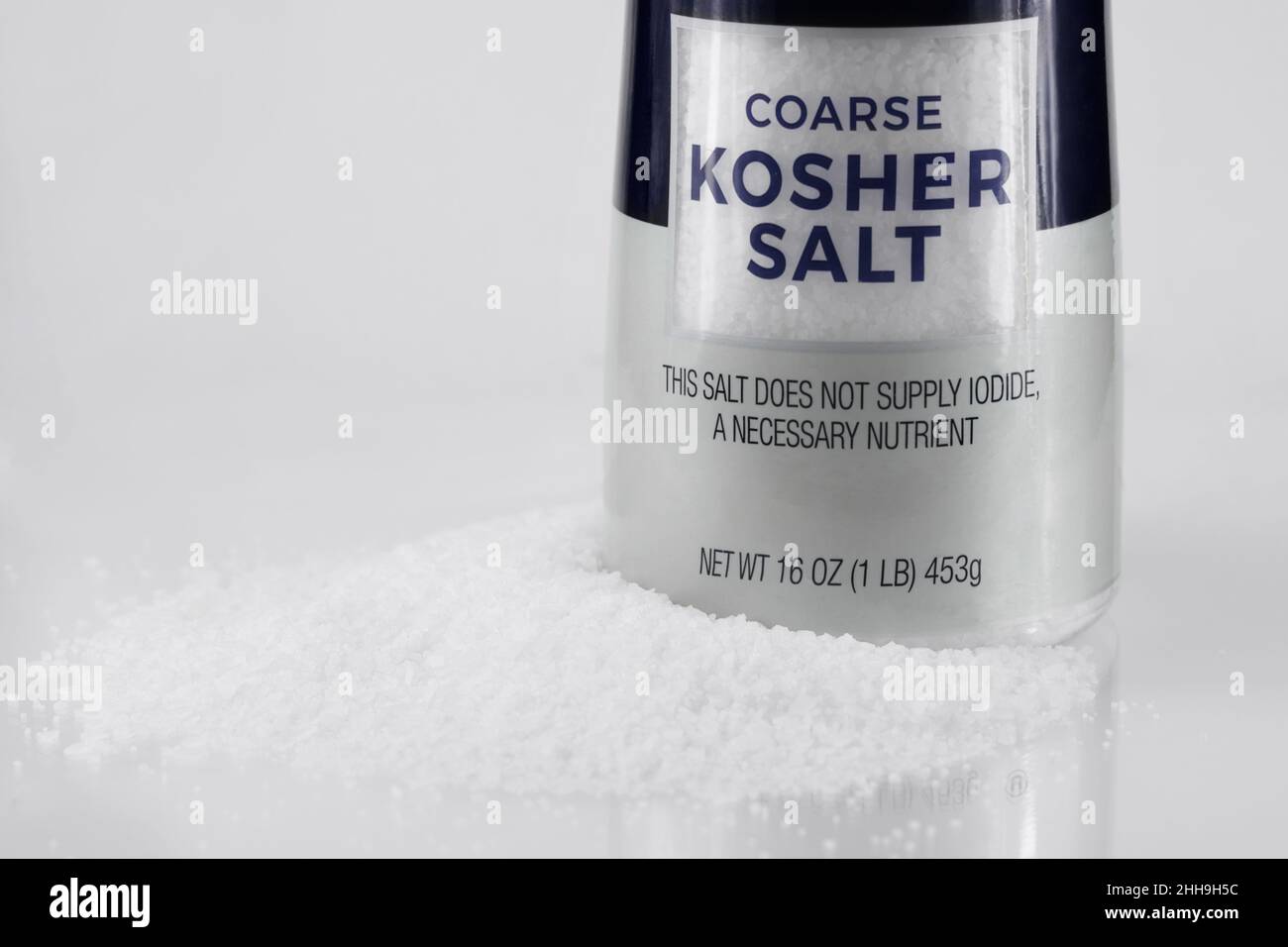 una botella de sal kosher gruesa rodeada por un charco de granos de sal sobre un fondo blanco, con espacio de copia Foto de stock