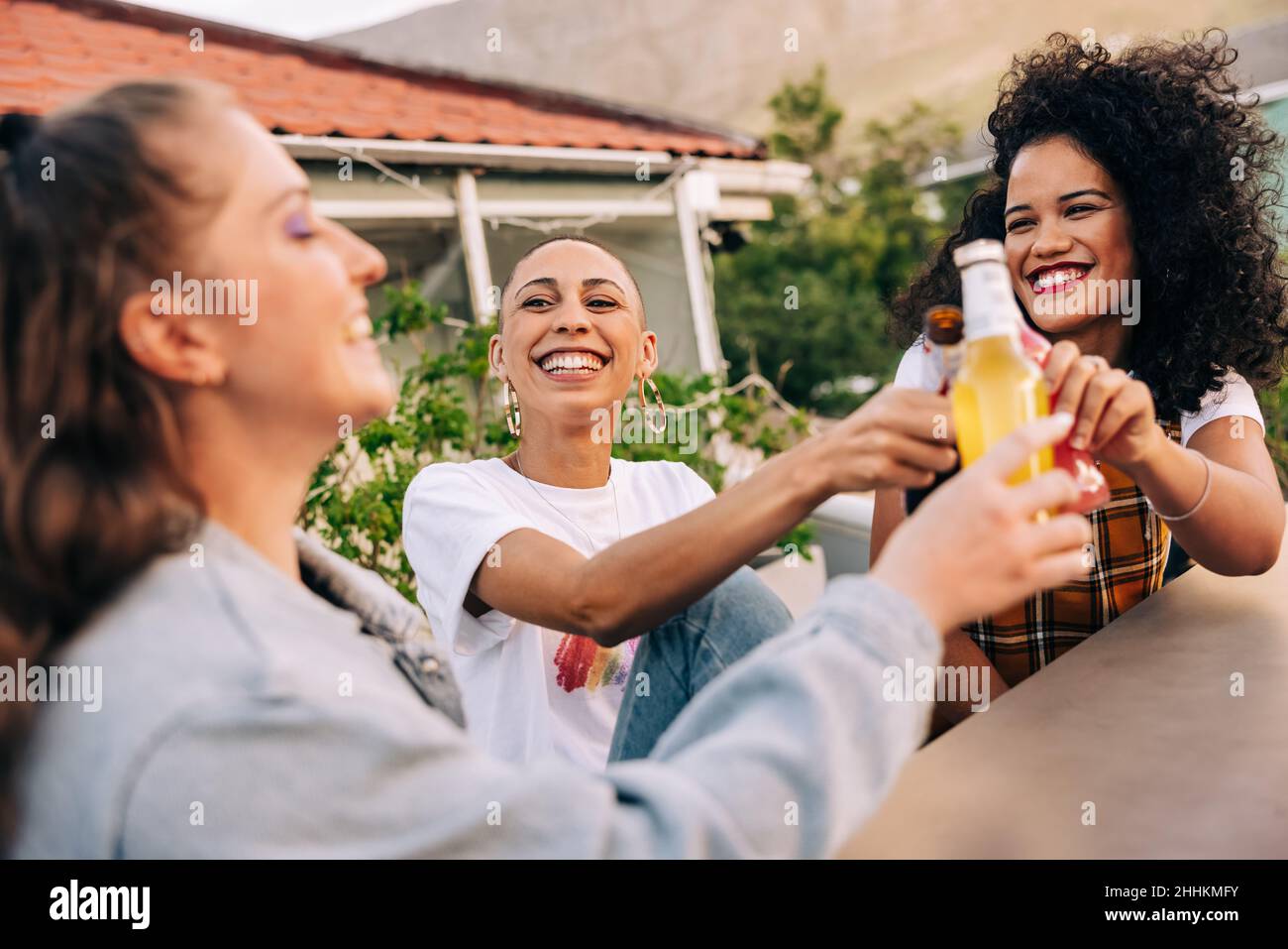 Novias haciendo un brindis a su amistad en un tejado. Tres amigas sonriendo alegremente mientras levantan sus botellas de cerveza. Amigos felices h Foto de stock