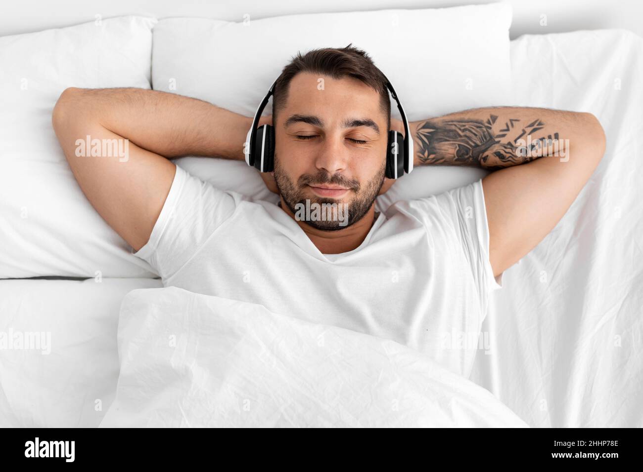 Satisfecho tranquilo calma joven europeo musculoso con los ojos cerrados en los auriculares se encuentra en la cama blanca Foto de stock