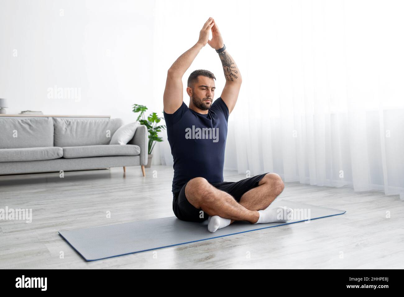 Calmado millennial musculoso caucásico masculino con ojos cerrados se sienta en el piso y practica yoga en el interior de la sala de estar Foto de stock
