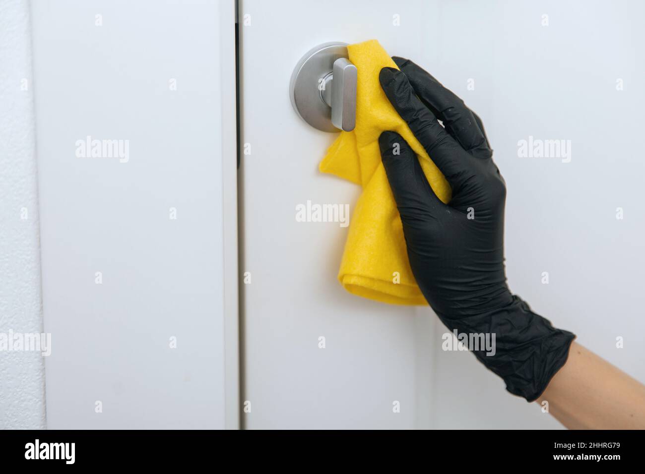 Limpieza del tirador de la puerta con un paño amarillo en guantes negros. Mano de mujer usando toalla para la limpieza. Desinfección en hospitales y espacios públicos contra corona Foto de stock