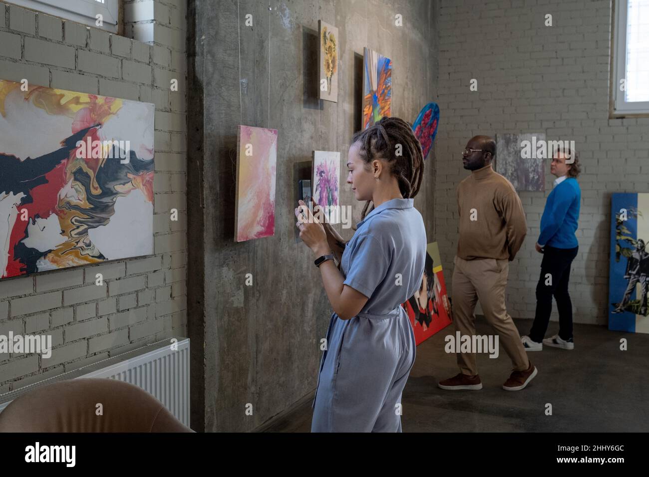 Joven fotografiando las pinturas en su teléfono móvil durante su visita a la galería de arte con otras personas en el fondo Foto de stock