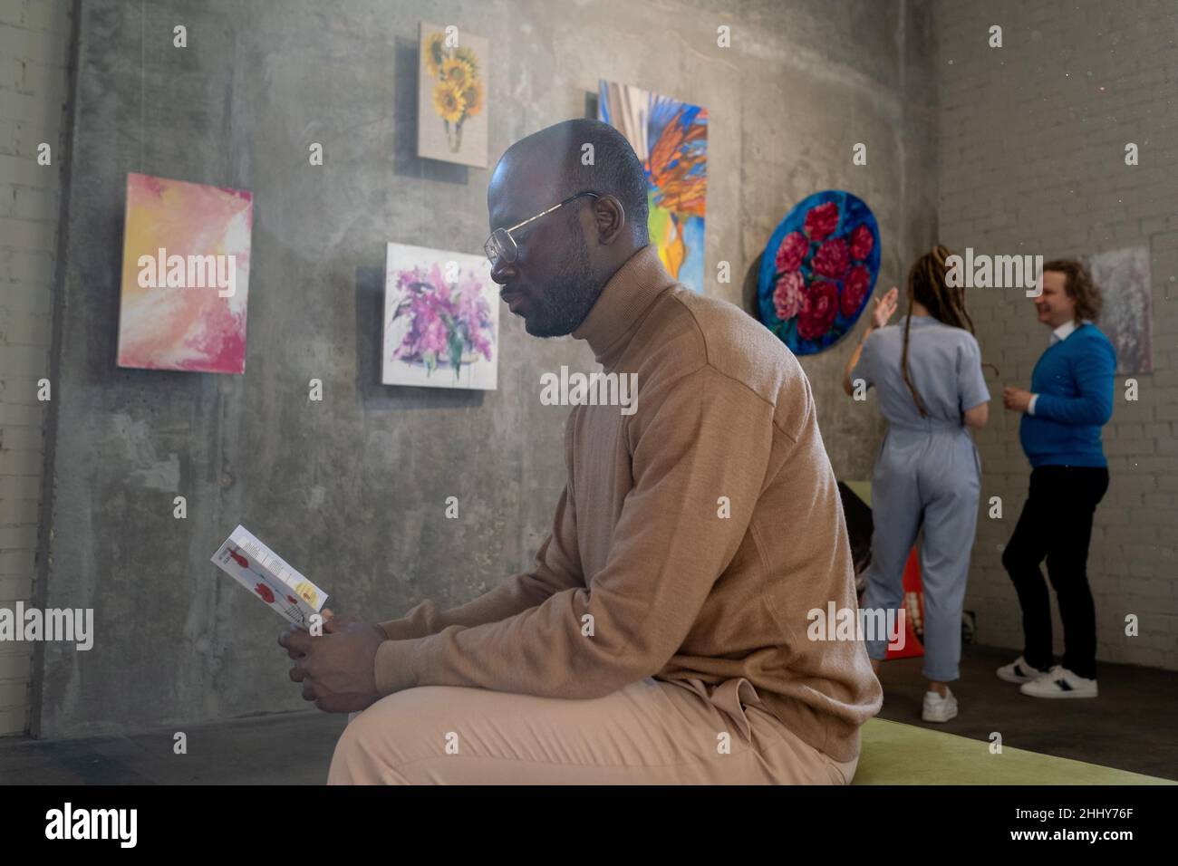 Un hombre serio africano con gafas leyendo un folleto mientras está sentado en la galería de arte con otras personas de fondo Foto de stock