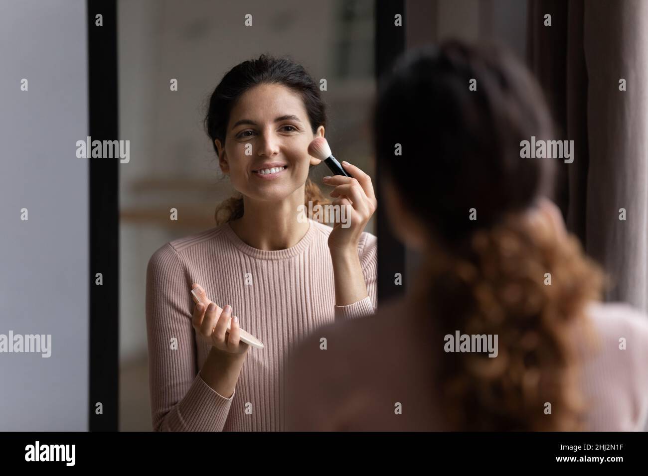 La mujer mira en el espejo sostiene el cepillo aplicando polvo sobre pómulos Foto de stock