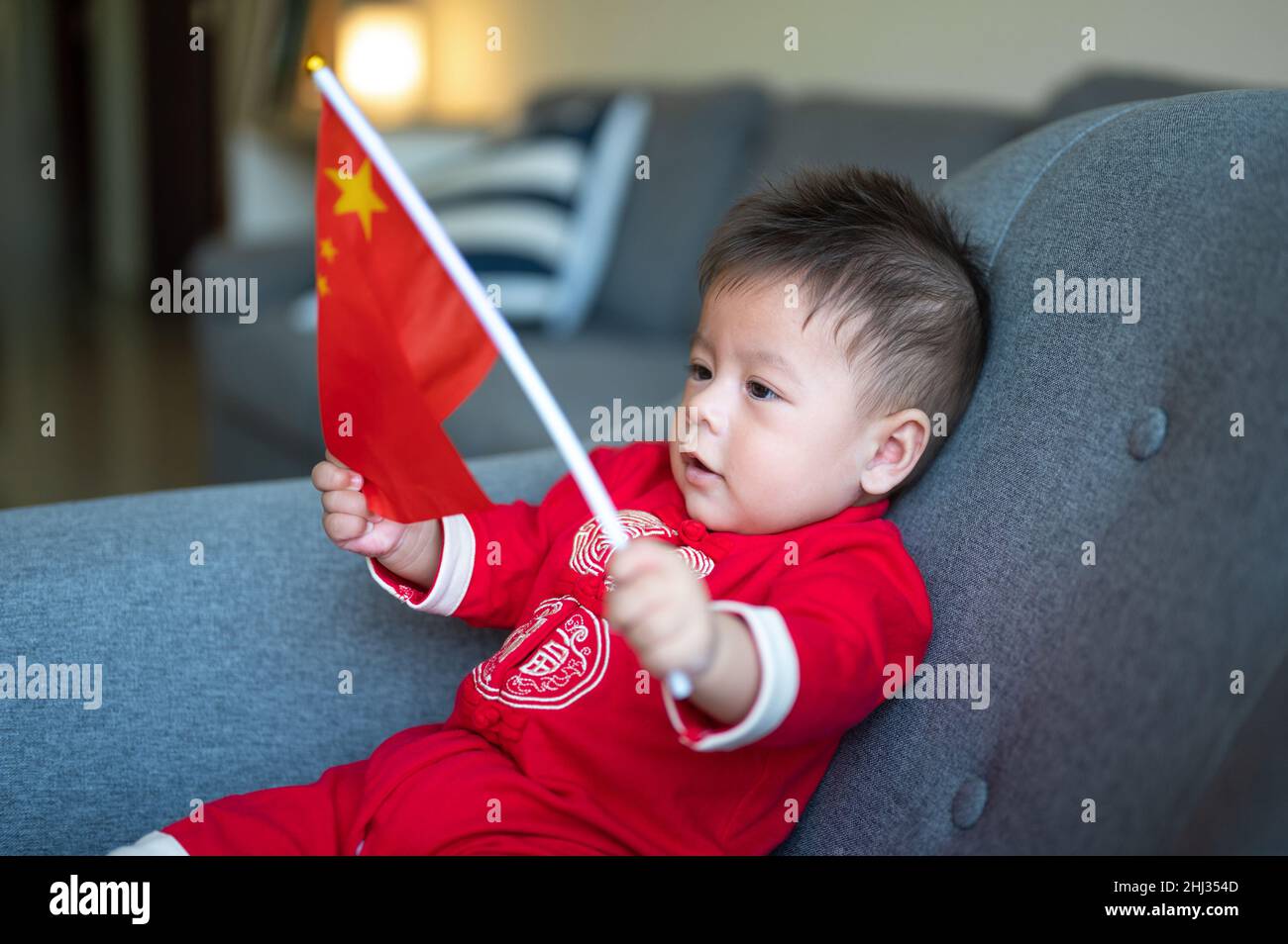 Niño asiático sosteniendo la bandera china sentado en un sofá mientras llevaba ropa tradicional china roja para el Año Nuevo Lunar en casa Foto de stock