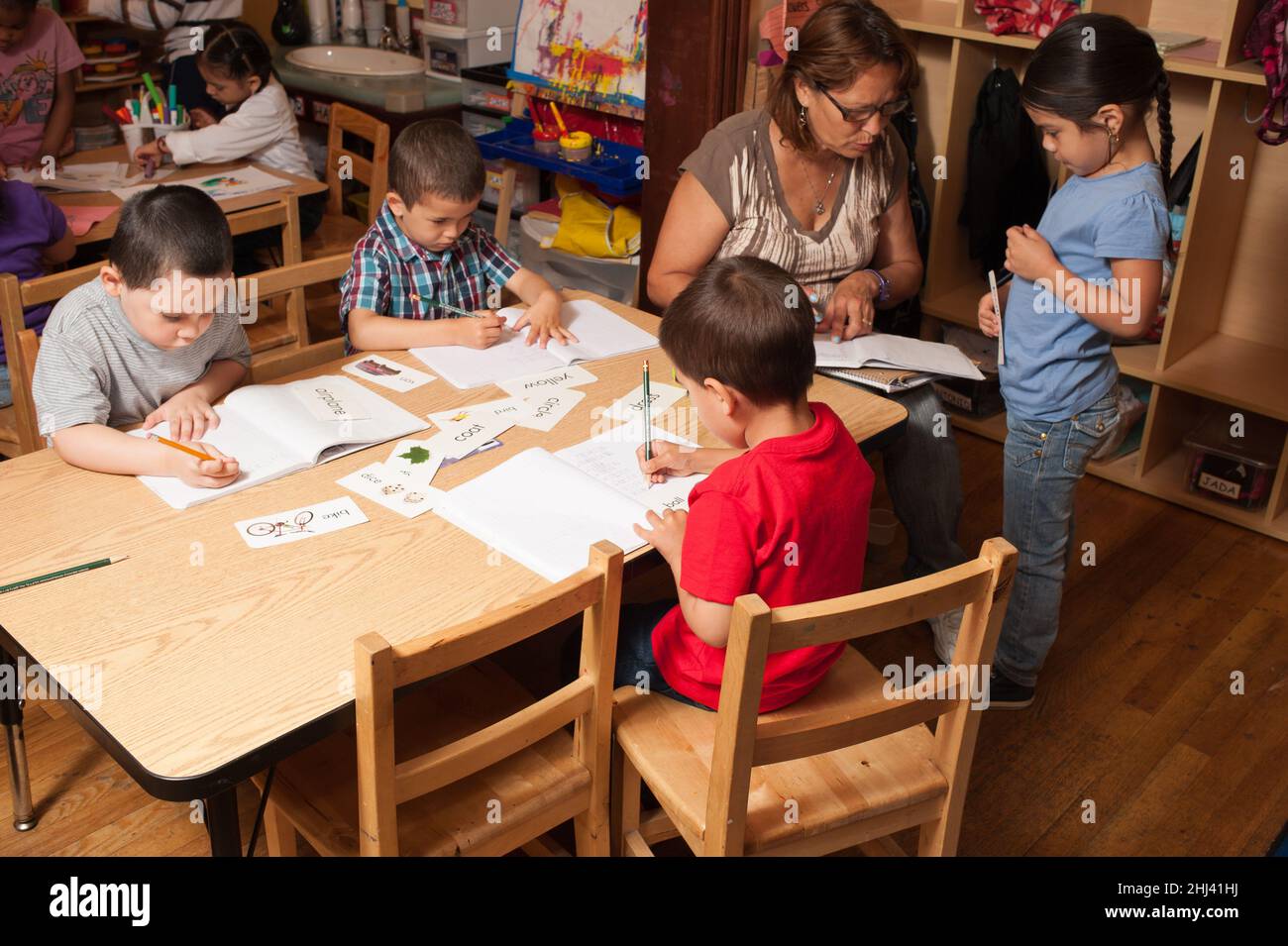 Educación Preescolar Head Start de 4-5 años de edad maestra mujer trabajando con niña mientras otros niños escriben en revistas con lápices Foto de stock