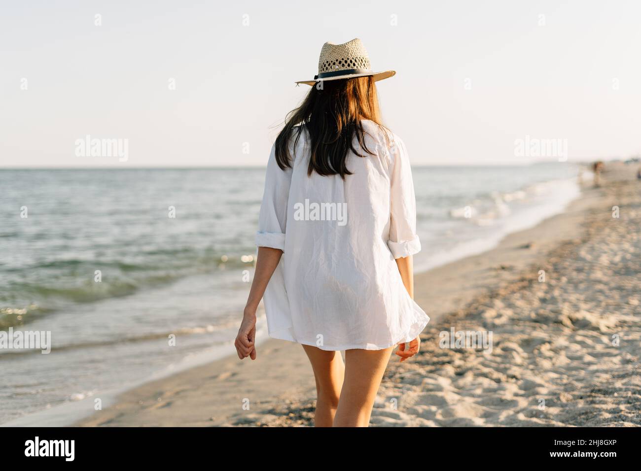Alrededor Artefacto Gama de La espalda de una mujer en una camisa y un sombrero camina en una playa  soleada con el mar azul en el fondo. El concepto de vacaciones y vacaciones  Fotografía de stock -