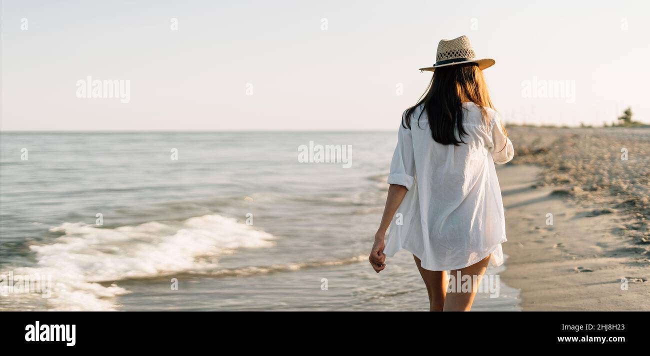 Vista trasera sin cara Feliz mujer camisa blanca disfruta de sus vacaciones tropicales en la playa paseos a lo largo del océano disfruta de la puesta de Fotografía de