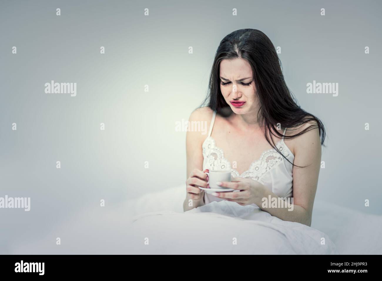 Una mujer se sienta en la cama y bebe café malo. La morena tiene una taza con una bebida caliente. Dormitorio blanco. Buenos días. Foto de stock