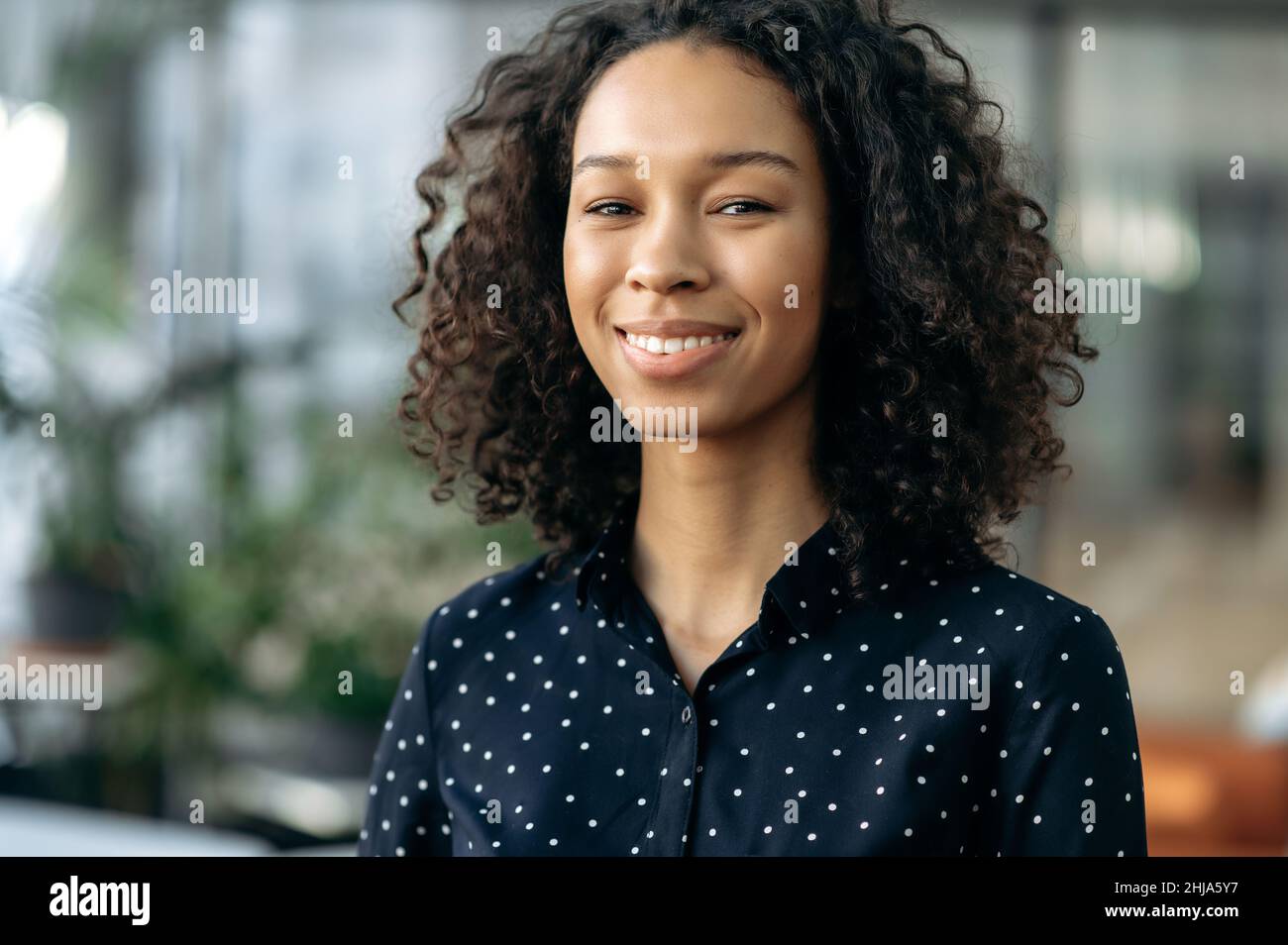 Primer plano de la curly atractiva niña afroamericana positiva, agradable joven de negocios, estudiante o freelancer, agente inmobiliario, de pie en el cargo, mirando la cámara, sonriendo amable Foto de stock