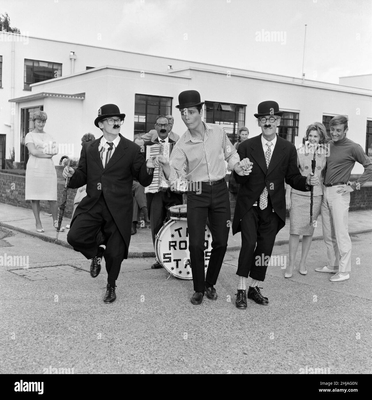 ¿Reconoce al 'busker' con la forma de un arquero? Es Cliff Richard, haciendo un juego de zapatos suaves con los buskers Road Stars de Londres. Cliff los había invitado a Elstree Studios donde está filmando vacaciones de verano para registrar su propia protesta por la reciente prohibición de los autobuses. Más tarde, los autobuses se fueron a ensayar para un compromiso en Westminster. Cliff preguntó: '¿No tienes miedo de quedar atrapado por la obstrucción?' Dijo los viejos 'uns al joven: 'No es una oportunidad. Estamos actuando en la boda de un policía.' Agosto de 1962. Foto de stock