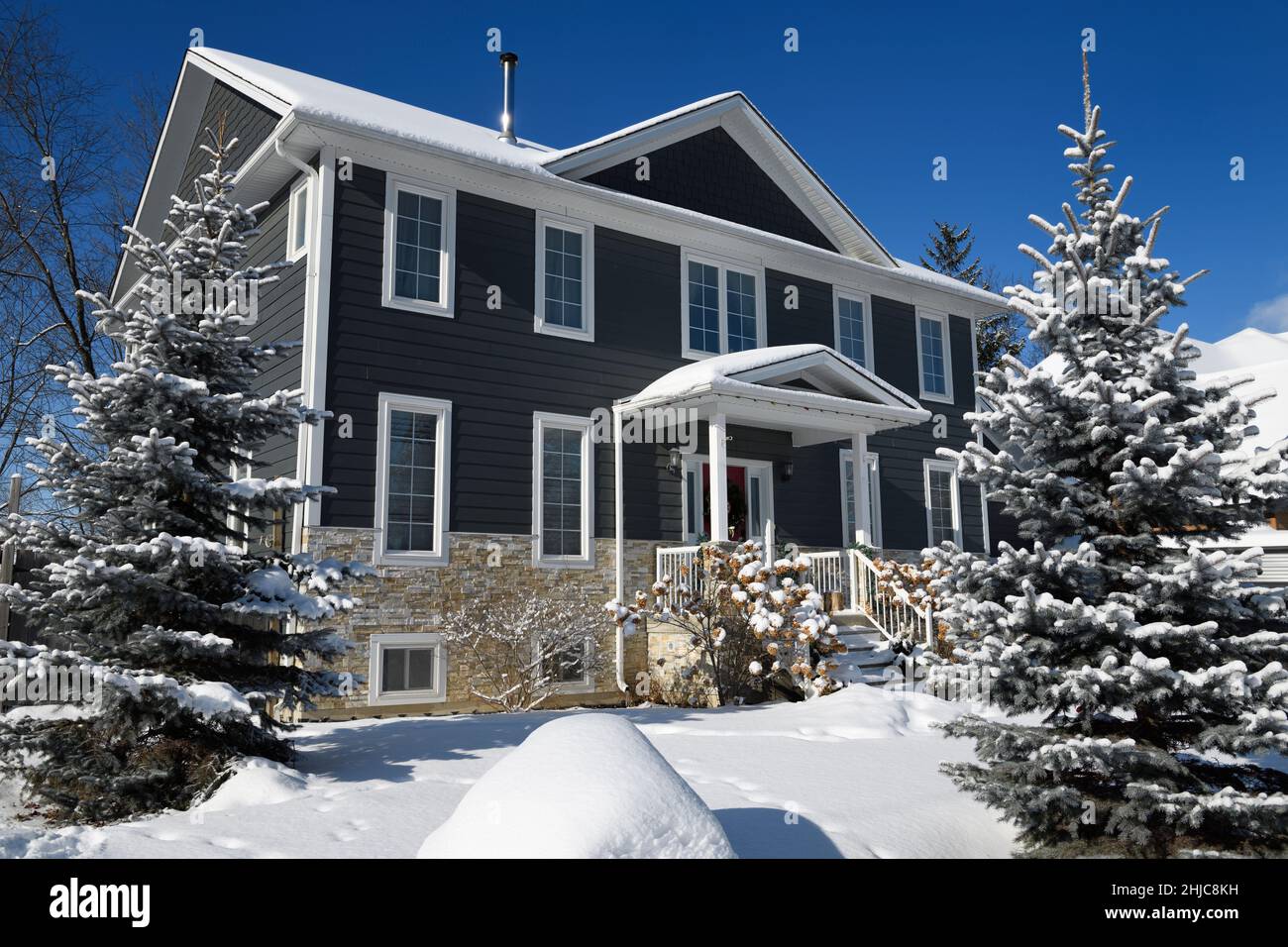 Sol sobre nieve fresca en árboles perennes y casa gris en Ontario Canadá invierno con cielo azul Foto de stock