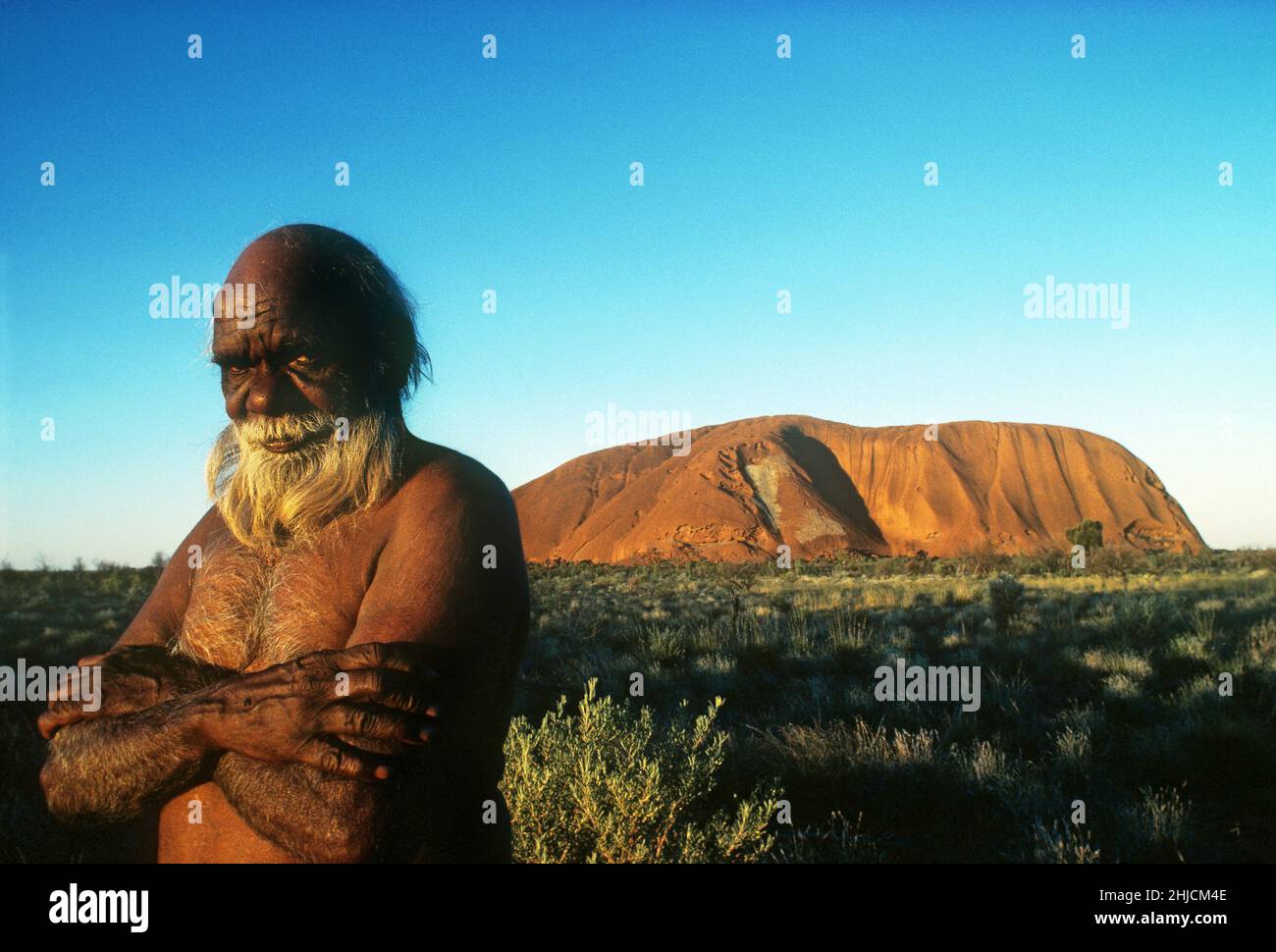 Toby Kaninga, un australiano indígena, se encuentra cerca de la gran formación de arenisca conocida como Ayers Rock. El monumento es sagrado para los aborígenes de la zona, lo que a veces convierte al turismo en una cuestión delicada. Foto c. 1982. Foto de stock