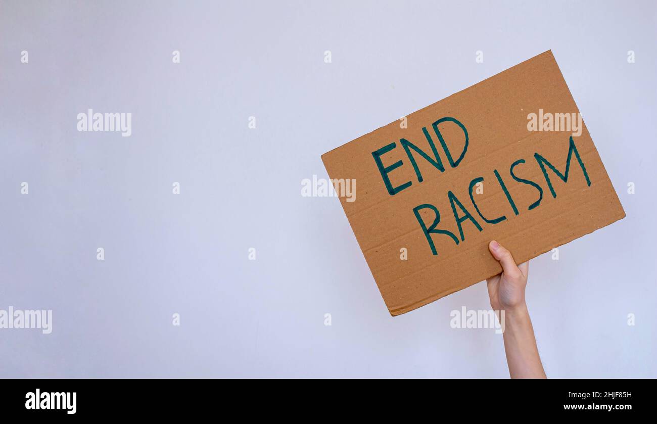 Mano sosteniendo cartón con inscripción 'End racismo' sobre fondo de pared gris. La mujer está protestando contra el racismo. Foto de stock