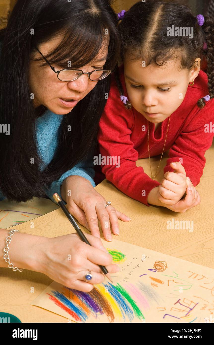 Salón de clases preescolar de 4-5 años, maestra con niña diciendo en voz alta lo que está escribiendo para su niño en su dibujo Foto de stock