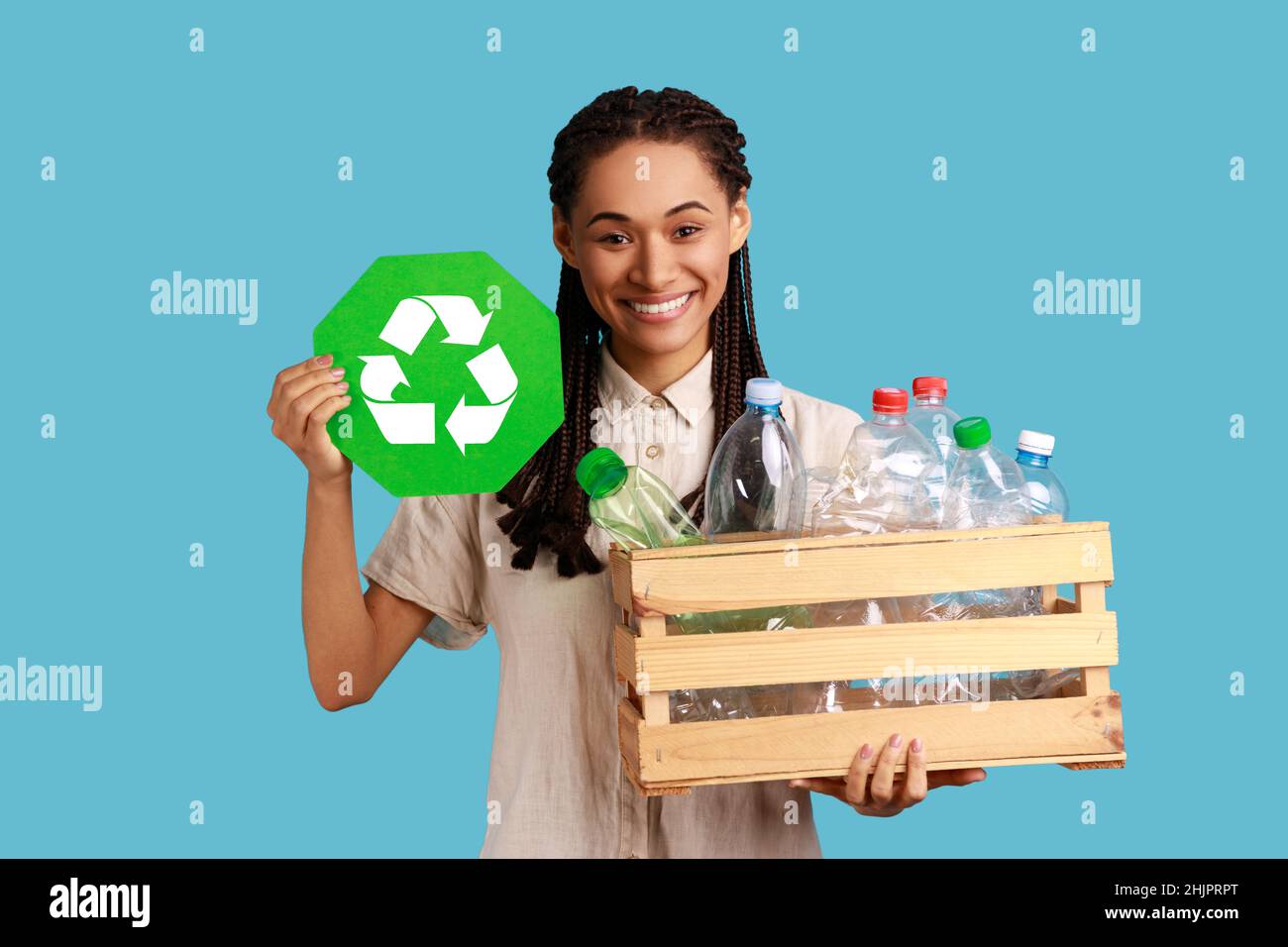 Mujer sonriente con dreadlocks negros caja de sujeción con botellas de plástico, reciclaje, clasificación de residuos y sostenibilidad, con camisa blanca. Estudio en interior grabado aislado sobre fondo azul. Foto de stock