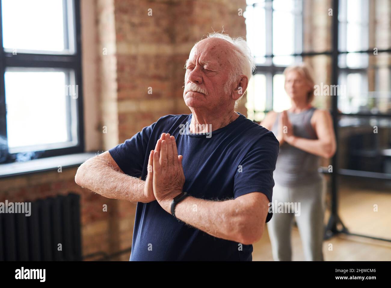 Hombre mayor de pie en yoga pose con sus ojos cerrados y meditando durante la clase con otras personas en el club de salud Foto de stock