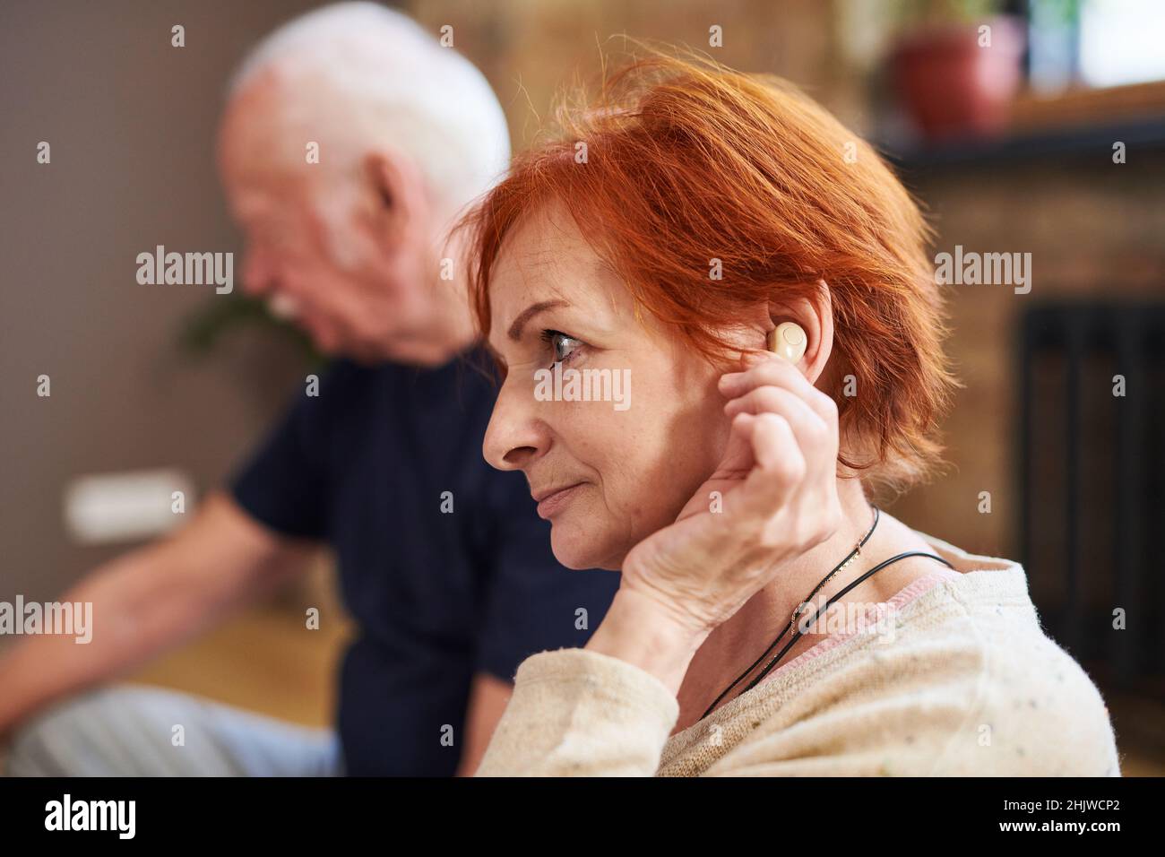 Vista lateral de una mujer mayor con el pelo rojo corto usando su audífono mientras se sienta con otras personas en clase de yoga Foto de stock