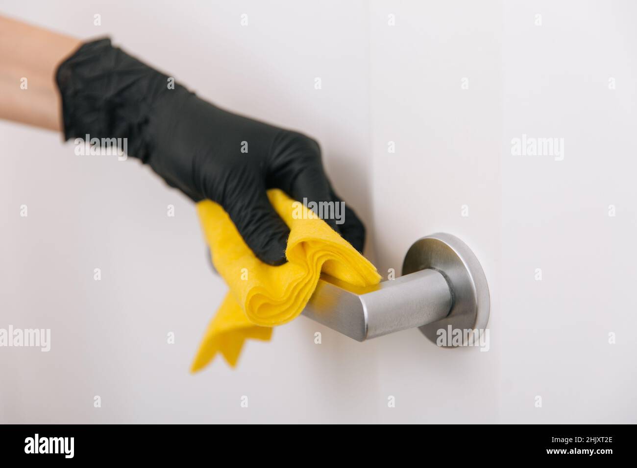 Limpieza del tirador de la puerta con un paño amarillo en guantes negros. Mano de mujer usando toalla para la limpieza. Desinfección en hospitales y espacios públicos contra corona Foto de stock