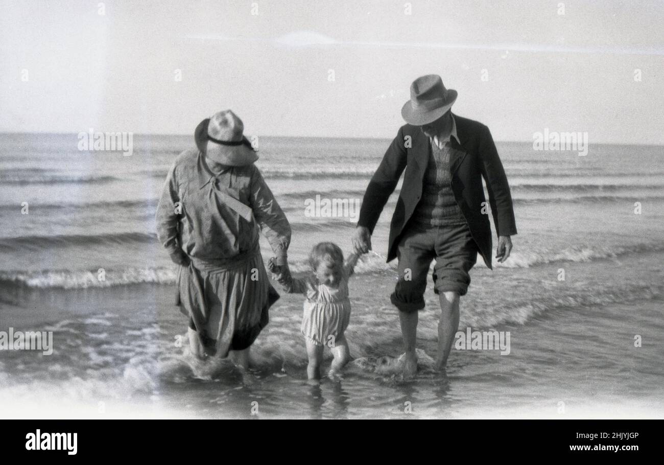 1930s, histórico, dos abuelos cada uno sosteniendo una mano de su nieta en las aguas poco profundas de la orilla del mar, Inglaterra, Reino Unido. La señora sostiene su vestido largo, que se ha mojado, mientras que los pantalones del caballero se enrollan hasta las rodillas. Aunque está en la costa, ambos llevan sombreros, típicos de la época. Foto de stock