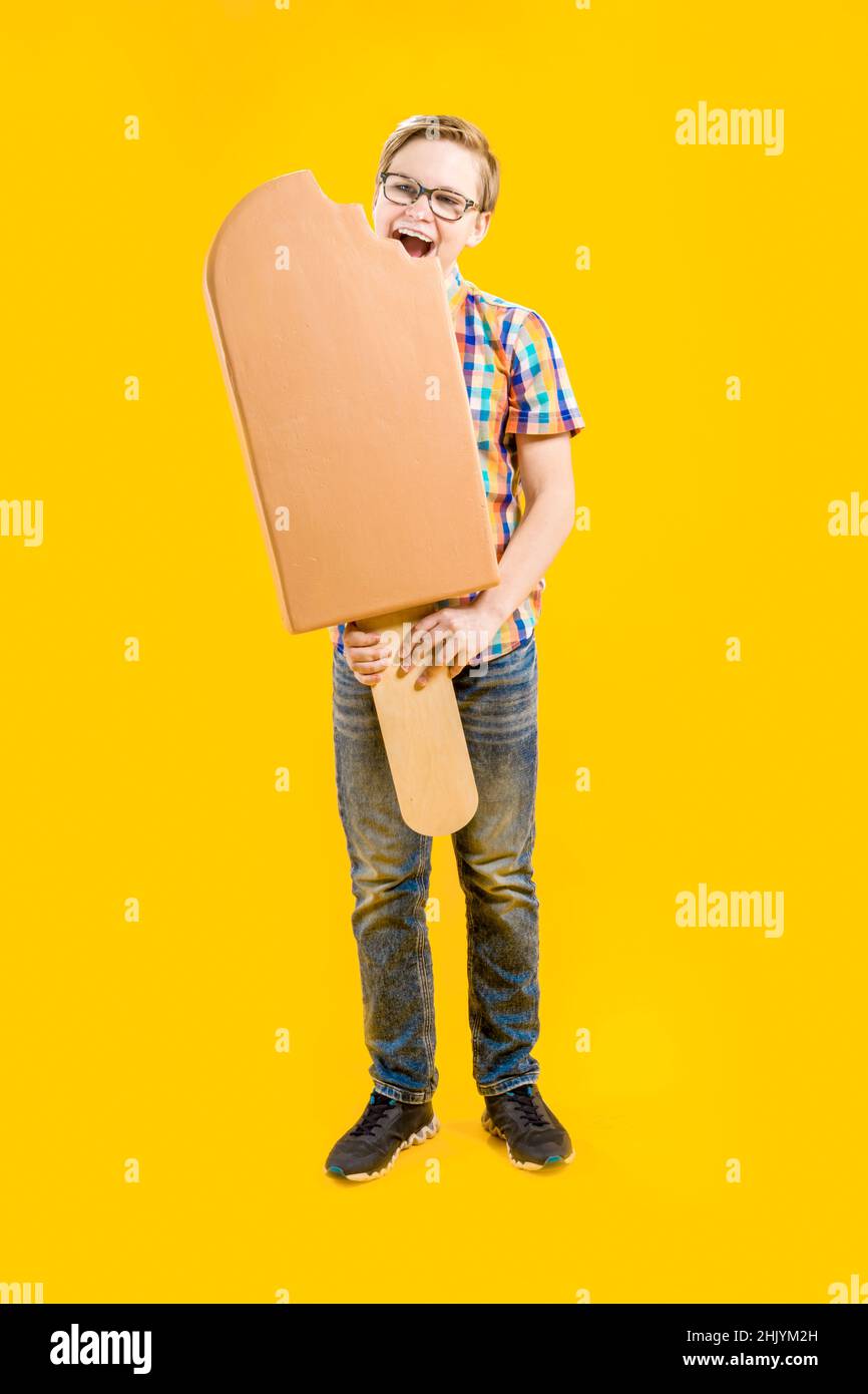 Un chico lindo está sosteniendo un enorme helado en un palo en sus manos, tratando de morderlo. Confitería. Delicioso postre. Adolescente alegre. Foto de stock