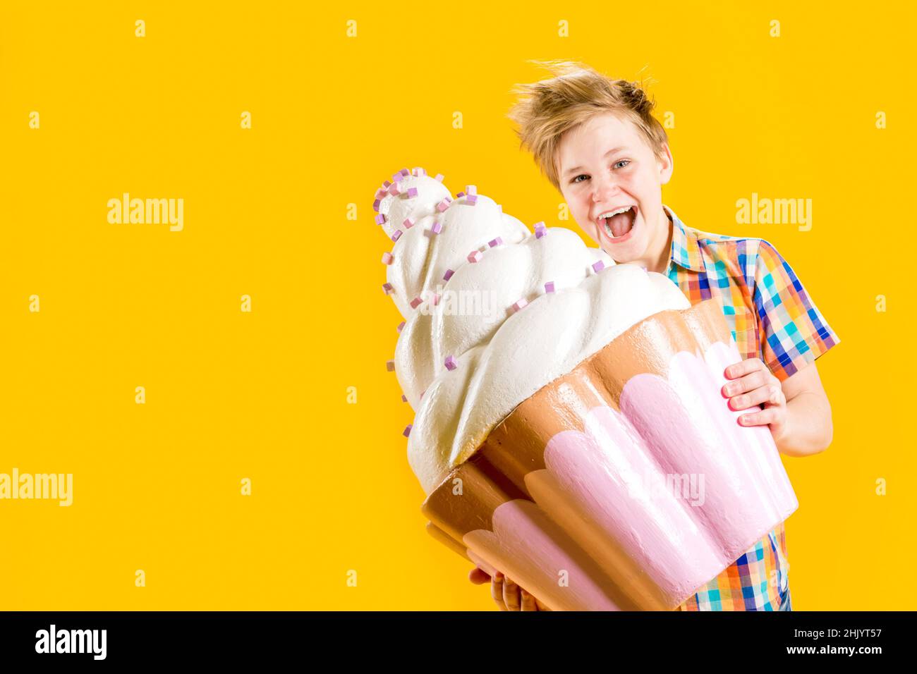 Un chico lindo sosteniendo un enorme cupcake en sus manos, tratando de morderlo sobre un fondo amarillo. Adolescente alegre con un corte de pelo descuidado. Confitería. D Foto de stock