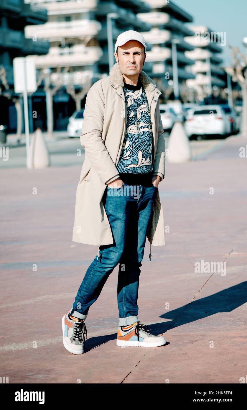 un hombre de mediana edad, con un abrigo beige, un jersey moderno y  colorido y un par de jeans, de pie al aire libre, en un espacio urbano, en  un soleado día