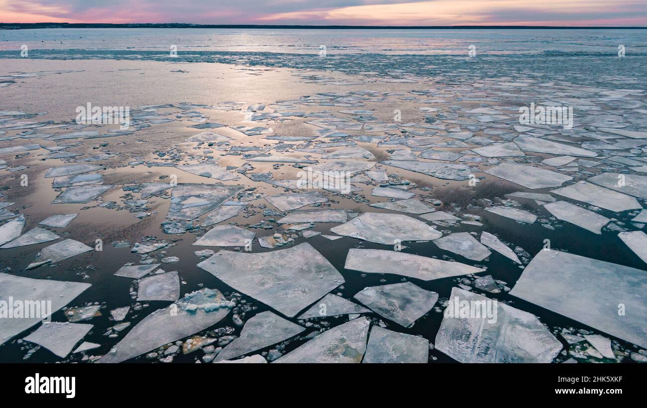 Pedazos de hielo gruesos rotos flotando en la superficie de aguas tranquilas cerca de la orilla del río en la brillante puesta de sol púrpura sobre el cielo nublado en la vista aérea de invierno Foto de stock