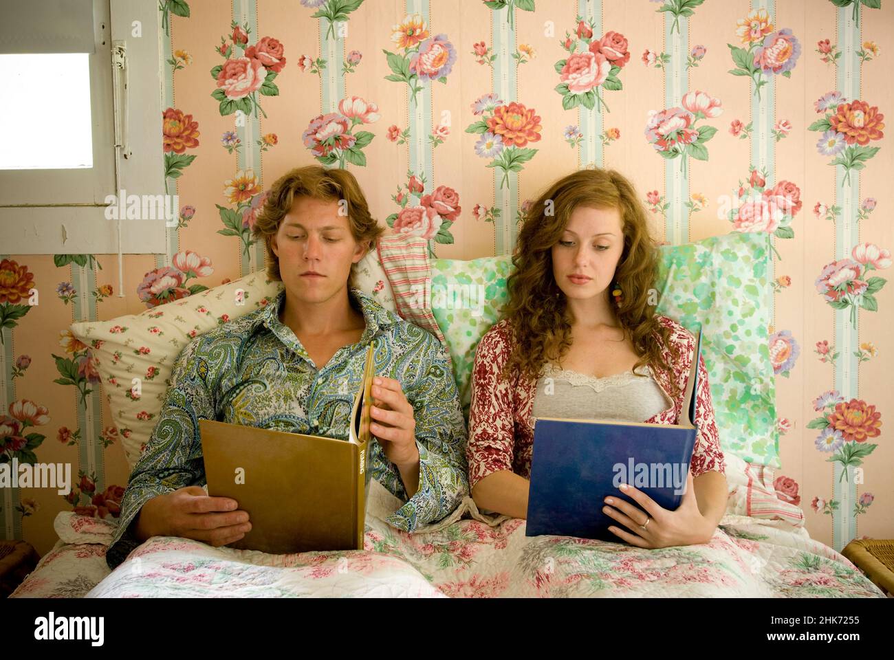 Una pareja joven tumbada en la cama leyendo libros en el dormitorio con paredes de papel pintado floral Foto de stock