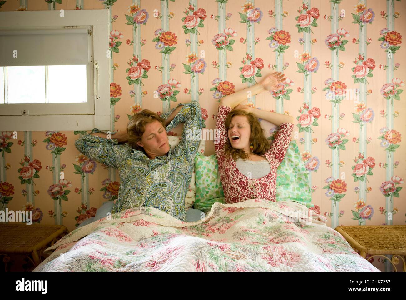 Pareja joven tumbada en la cama estirando y bostezando en el dormitorio con paredes de papel pintado floral Foto de stock