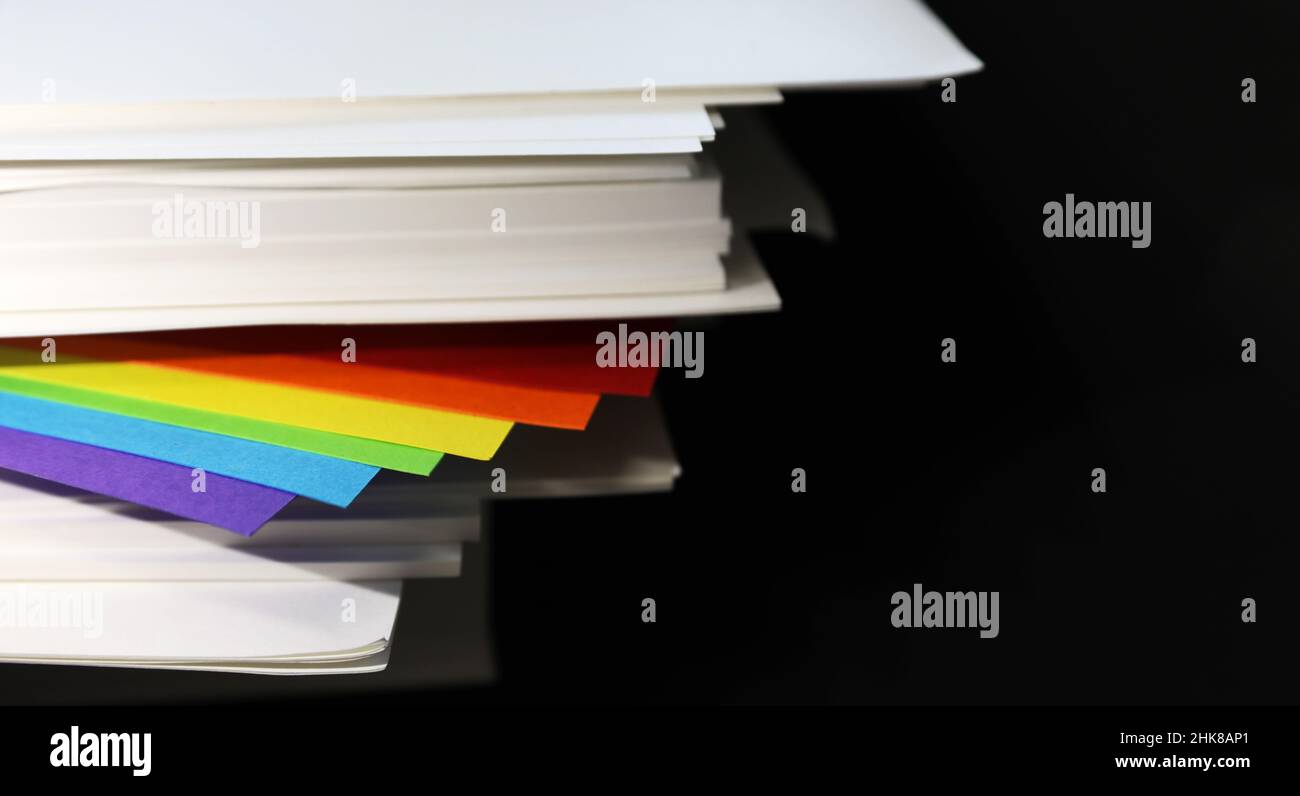 gay lesbiana bisexual queer transgender comunidad arcoiris bandera colores poking fuera de una resma de papel blanco crujiente de la oficina sobre un fondo negro liso. Foto de stock