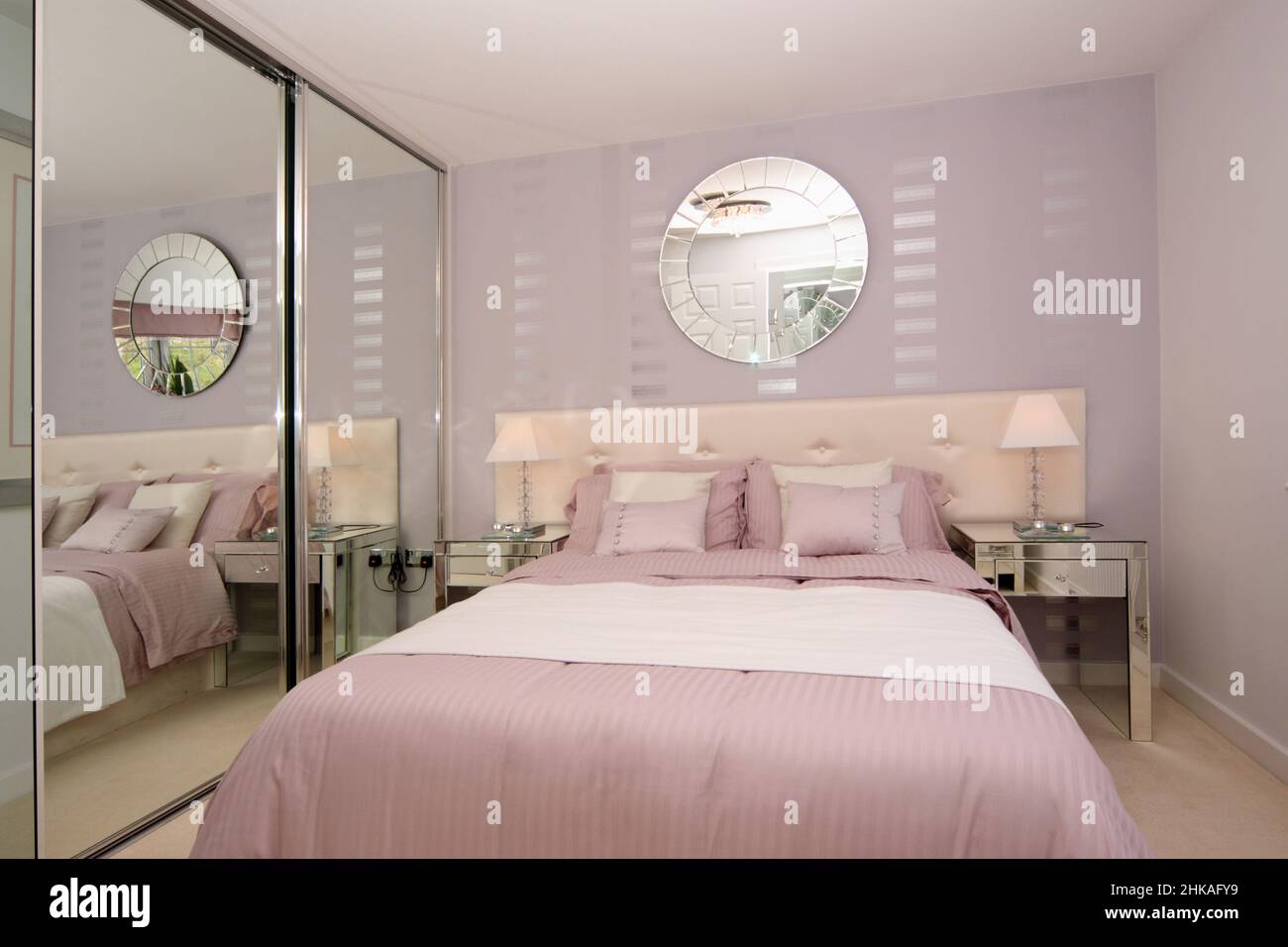 Dormitorio en combinación de colores rosa y blanco, con pared, luces de cabecera, cojines, espejo en la pared, cabecero acolchado, armarios con espejo Foto de stock