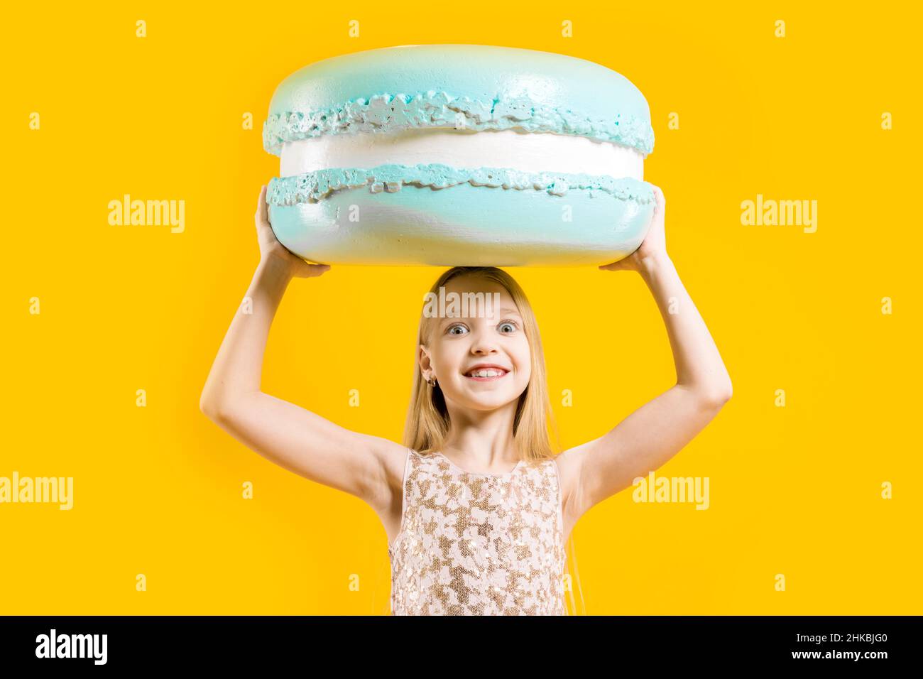 Una chica linda sostiene una enorme macaroons azul sobre su cabeza sobre un fondo amarillo. Regalo dulce. Postre redondo. Dulzura de Navidad. Foto de stock