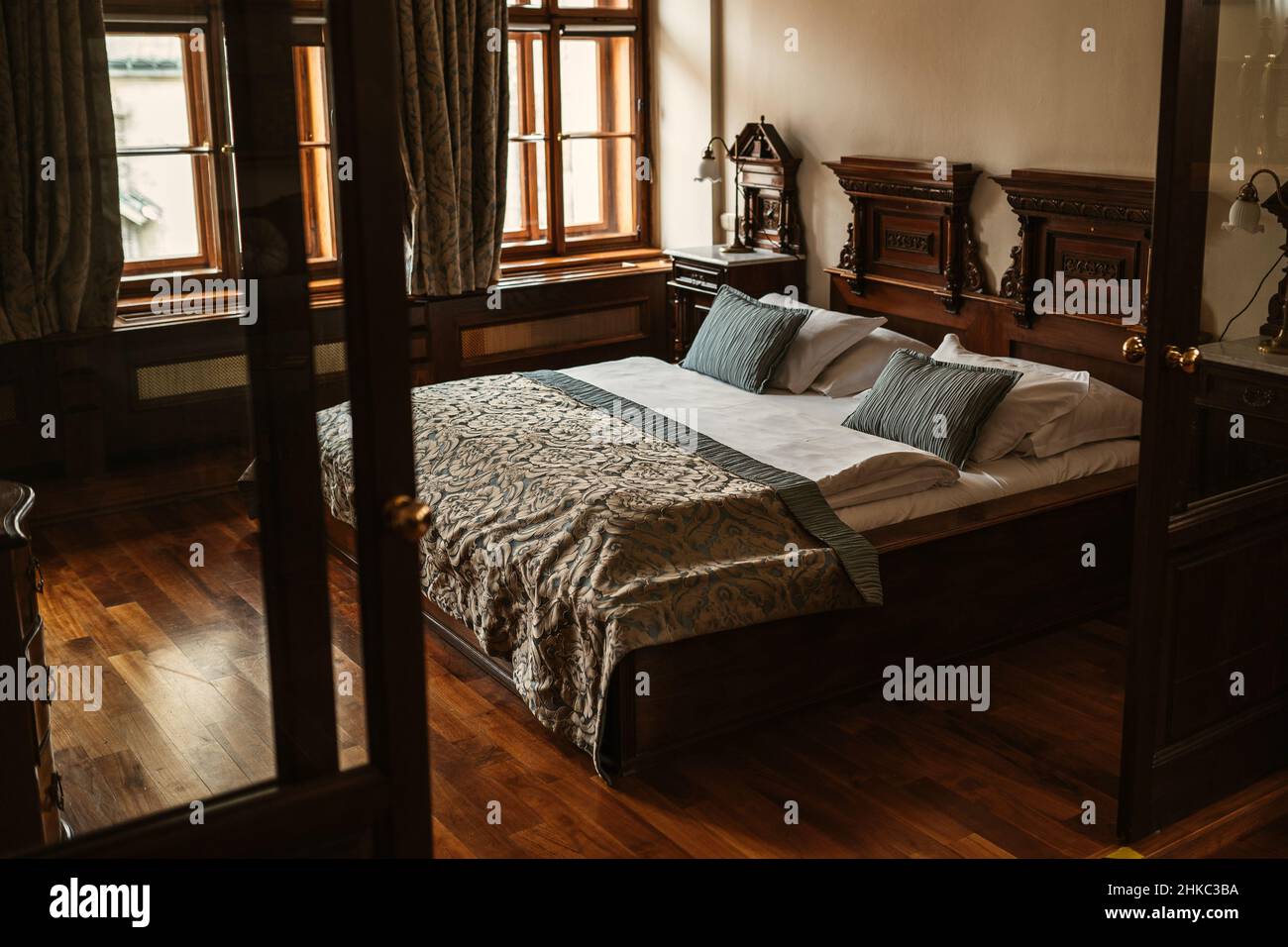 Elegante habitación medieval de lujo europeo con suelo de madera y cama con almohadas verdes, sábanas, colcha y lámpara de habitación en el fondo. Foto de stock