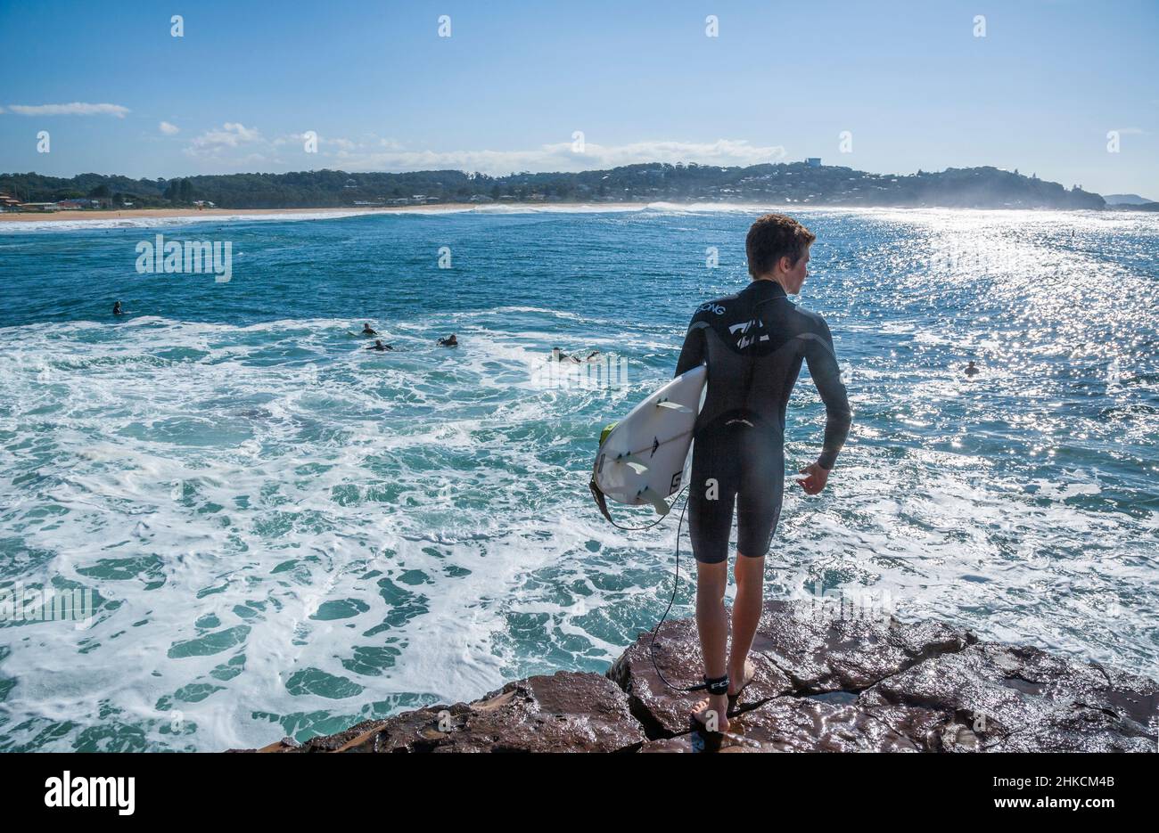 Joven surfista contemplando entrar en el tumultuoso surf desde la peligrosa plataforma de roca costera en Mups Rock, Avoca Beach, Central Coast, New South WA Foto de stock