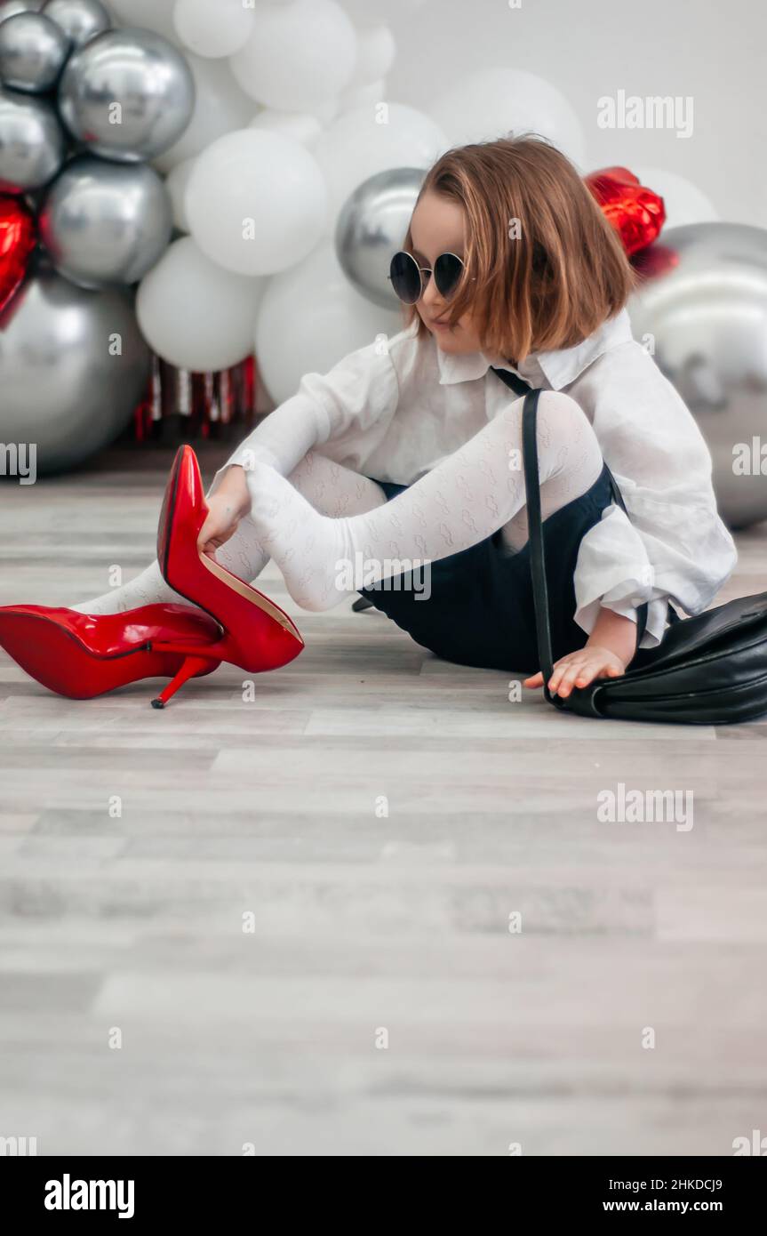 Un bebé de moda con gafas de sol está tratando con los zapatos rojos de su  madre. Moda y ropa moderna. Brillo brillante fondo de espejo metálico,  nuevas tendencias v Fotografía de