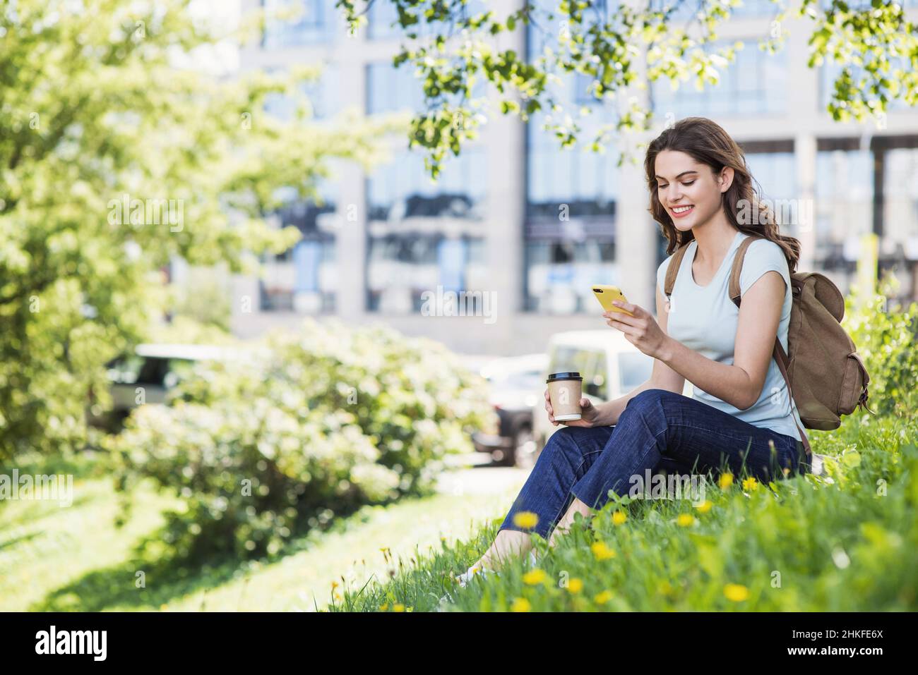 Mujer sonriente usando el teléfono inteligente en un parque de la ciudad. Joven hermosa niña teniendo café romper. Estilo de vida del estudiante, concepto de conexión Foto de stock