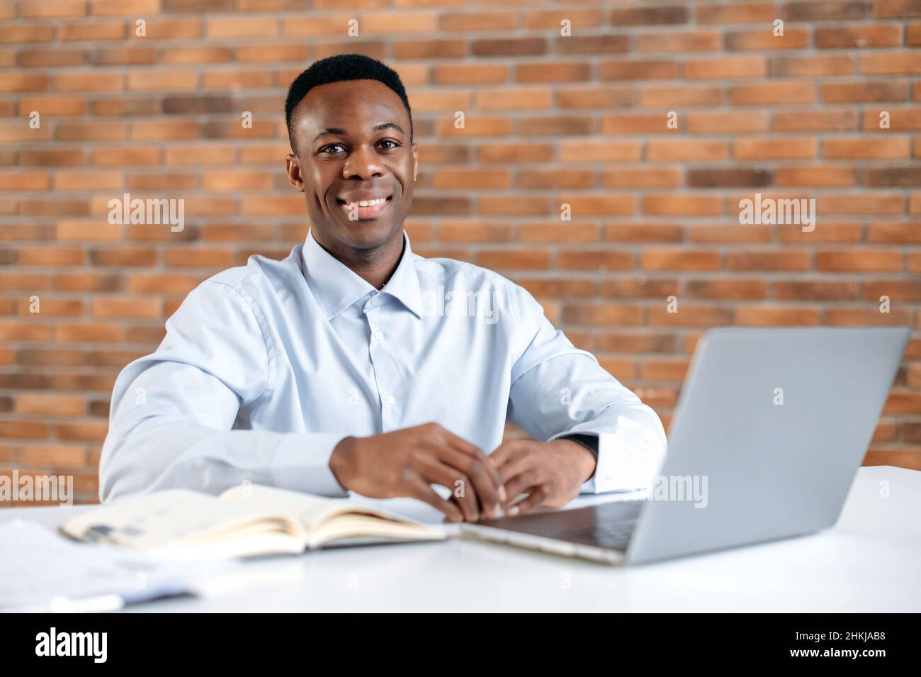 Retrato de un atractivo afroamericano en la oficina moderna. Agradable hombre inteligente de éxito, gerente superior de la empresa o comerciante, trabajando con el portátil en el lugar de trabajo, mira la cámara, sonreír amable Foto de stock