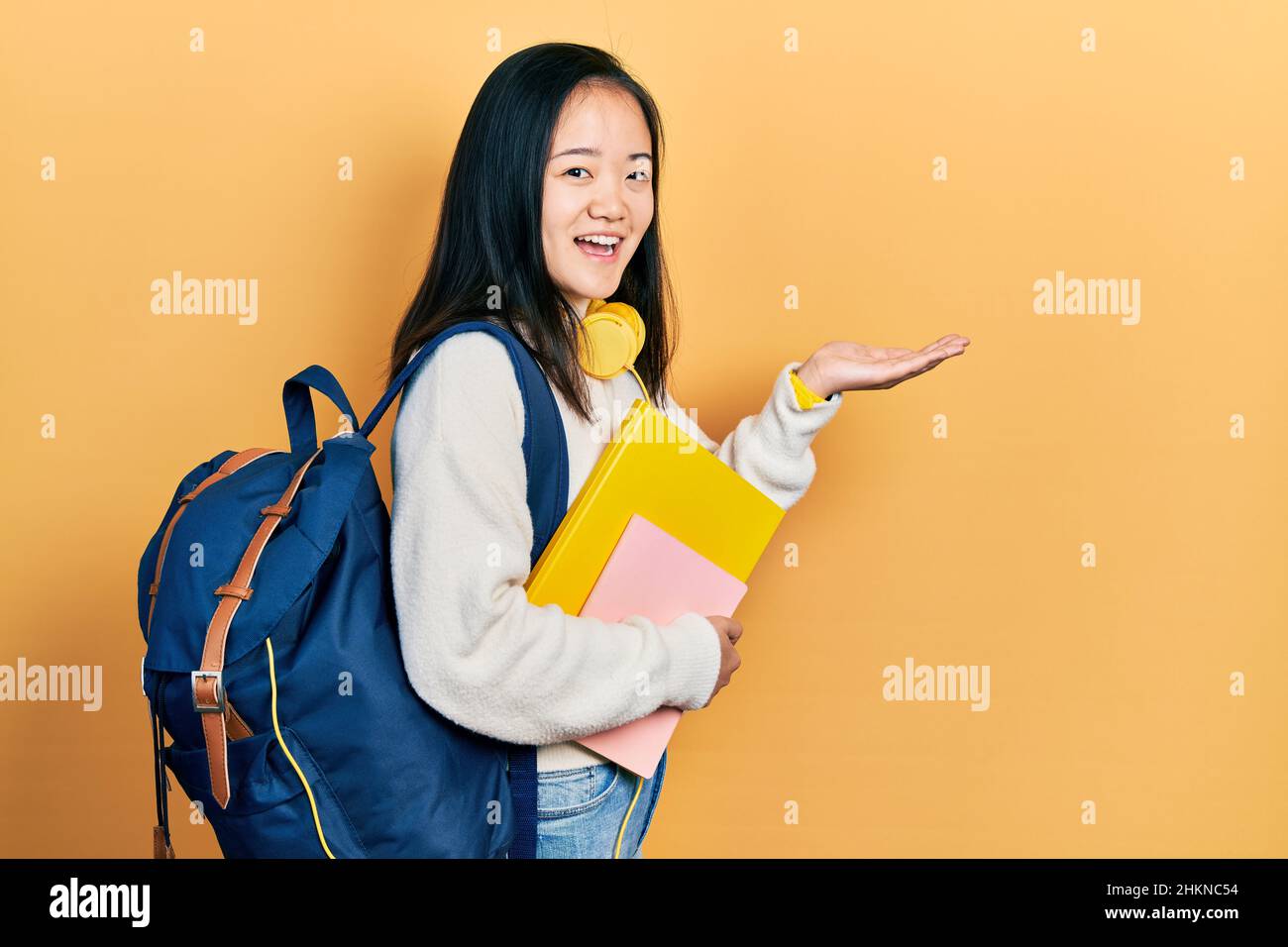 Joven china sosteniendo la mochila del estudiante y los libros señalando a  un lado con las manos abiertas palmeras que muestran espacio de copia,  presentando anuncio sonriendo excite Fotografía de stock -