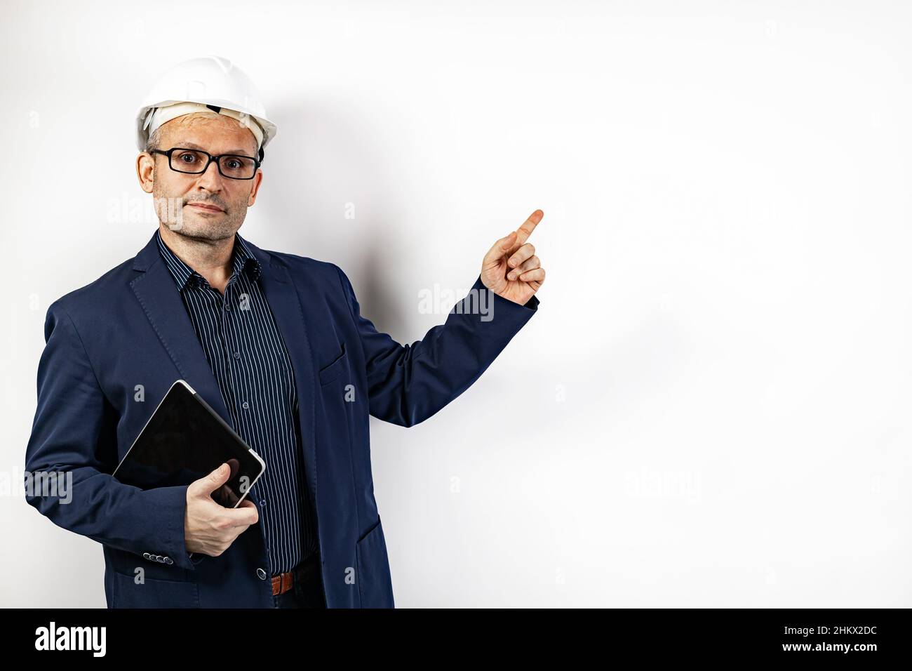 Un ingeniero masculino en un casco duro blanco apunta un dedo sobre un espacio de copia. Hombre de mediana edad con una chaqueta azul con un tablet. Retrato aislado activado Foto de stock