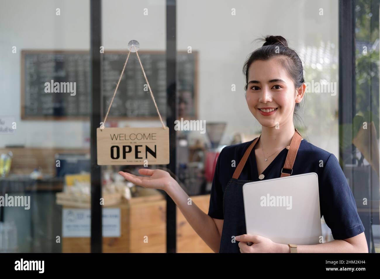 Hermosa mujer asiática joven barista en el delantal que sostiene la tableta y de pie en frente de la puerta de la cafetería con letrero abierto. PYME de inicio del propietario de la empresa Foto de stock