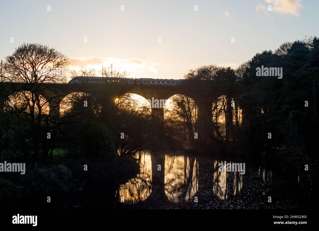El sol mira a través de un arco de un viaducto ferroviario al atardecer mientras un tren acelera por encima Foto de stock