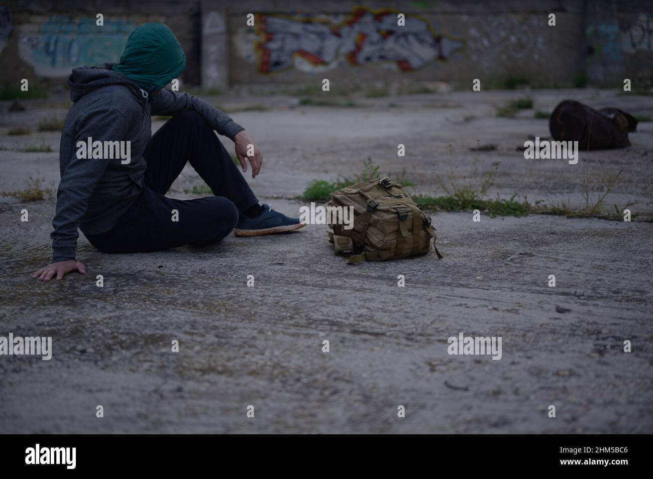 hombre sin hogar en hoodie sentado cerca de la mochila en asfalto Foto de stock