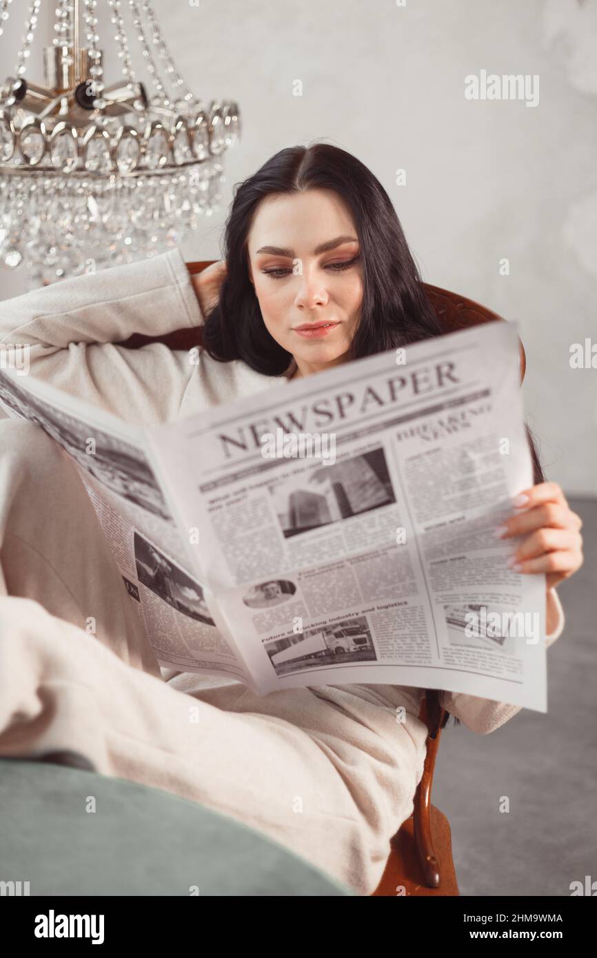 Una joven morena con ropa de casa lee el periódico con las piernas sobre la mesa. Concepto de fin de semana, lectura de noticias, prensa Foto de stock